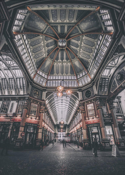 A wide-angle, symmetrical photograph looking down the center of an ornate Victorian covered market, Leadenhall Market. The focus is on the elaborate, arched glass and ironwork ceiling which is decorated in dark blue with gold stars. Below, a cobblestone path is lined with traditional shops with glowing lights, and a few people can be seen walking in the distance. The image has a moody, high-contrast aesthetic. Wall Art