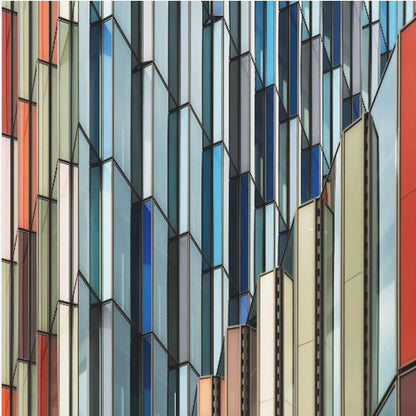 A close-up abstract photograph of a modern building's facade, featuring a complex pattern of colorful, angular glass panels in shades of blue, orange, red, and gray, all presented within a light wood frame. Wall Art
