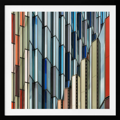 A close-up abstract photograph of a modern building's facade, featuring a complex pattern of colorful, angular glass panels in shades of blue, orange, red, and gray, all presented within a light wood frame. Wall Art