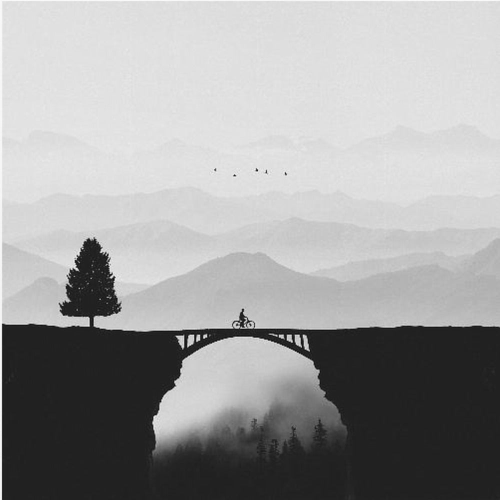 A minimalist black and white artwork featuring the silhouette of a cyclist crossing a high arched bridge between two cliffs. A lone tree stands on one cliff, while layers of misty mountains fill the background, creating a sense of depth and solitude. Poster
