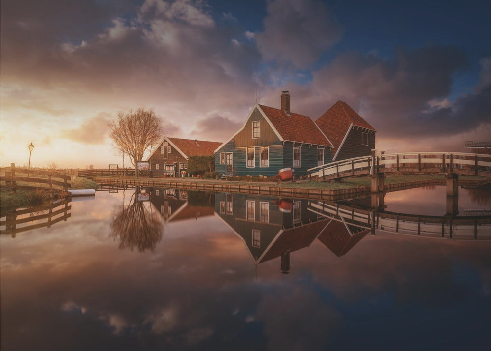 A picturesque landscape of traditional Dutch houses with teal walls and red roofs reflected perfectly in the calm water of a canal at sunset, under a dramatic, cloudy sky. A small wooden bridge crosses the canal, and the scene is enclosed in a silver frame. Artwork