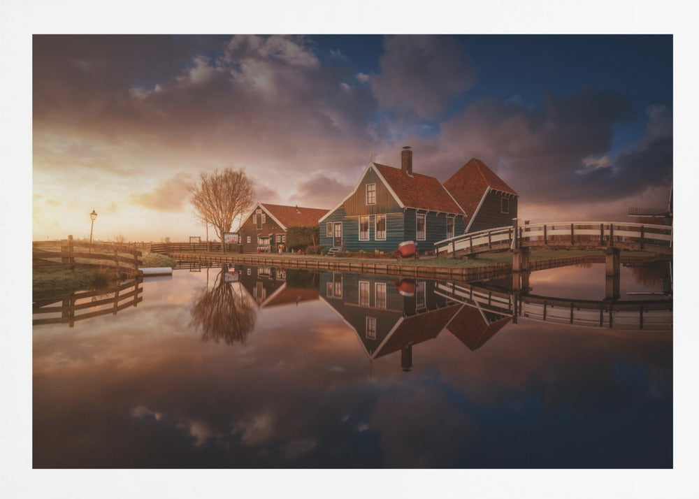 A picturesque landscape of traditional Dutch houses with teal walls and red roofs reflected perfectly in the calm water of a canal at sunset, under a dramatic, cloudy sky. A small wooden bridge crosses the canal, and the scene is enclosed in a silver frame. Artwork