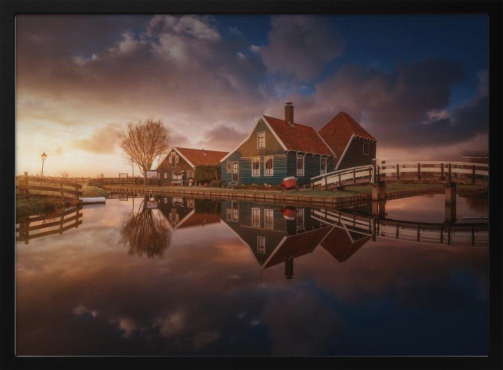 A picturesque landscape of traditional Dutch houses with teal walls and red roofs reflected perfectly in the calm water of a canal at sunset, under a dramatic, cloudy sky. A small wooden bridge crosses the canal, and the scene is enclosed in a silver frame. Artwork