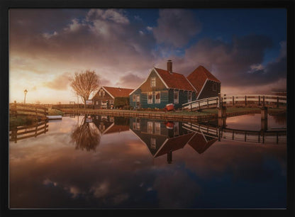 A picturesque landscape of traditional Dutch houses with teal walls and red roofs reflected perfectly in the calm water of a canal at sunset, under a dramatic, cloudy sky. A small wooden bridge crosses the canal, and the scene is enclosed in a silver frame. Artwork