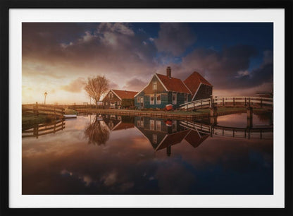 A picturesque landscape of traditional Dutch houses with teal walls and red roofs reflected perfectly in the calm water of a canal at sunset, under a dramatic, cloudy sky. A small wooden bridge crosses the canal, and the scene is enclosed in a silver frame. Artwork