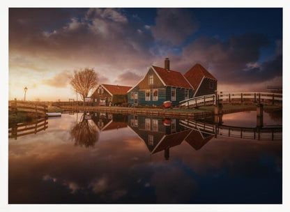 A picturesque landscape of traditional Dutch houses with teal walls and red roofs reflected perfectly in the calm water of a canal at sunset, under a dramatic, cloudy sky. A small wooden bridge crosses the canal, and the scene is enclosed in a silver frame. Artwork