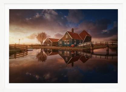 A picturesque landscape of traditional Dutch houses with teal walls and red roofs reflected perfectly in the calm water of a canal at sunset, under a dramatic, cloudy sky. A small wooden bridge crosses the canal, and the scene is enclosed in a silver frame. Artwork