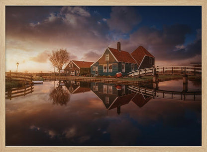 A picturesque landscape of traditional Dutch houses with teal walls and red roofs reflected perfectly in the calm water of a canal at sunset, under a dramatic, cloudy sky. A small wooden bridge crosses the canal, and the scene is enclosed in a silver frame. Artwork