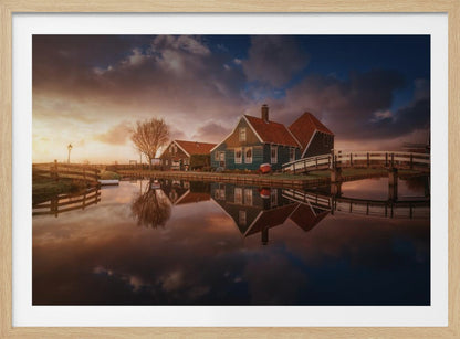 A picturesque landscape of traditional Dutch houses with teal walls and red roofs reflected perfectly in the calm water of a canal at sunset, under a dramatic, cloudy sky. A small wooden bridge crosses the canal, and the scene is enclosed in a silver frame. Artwork