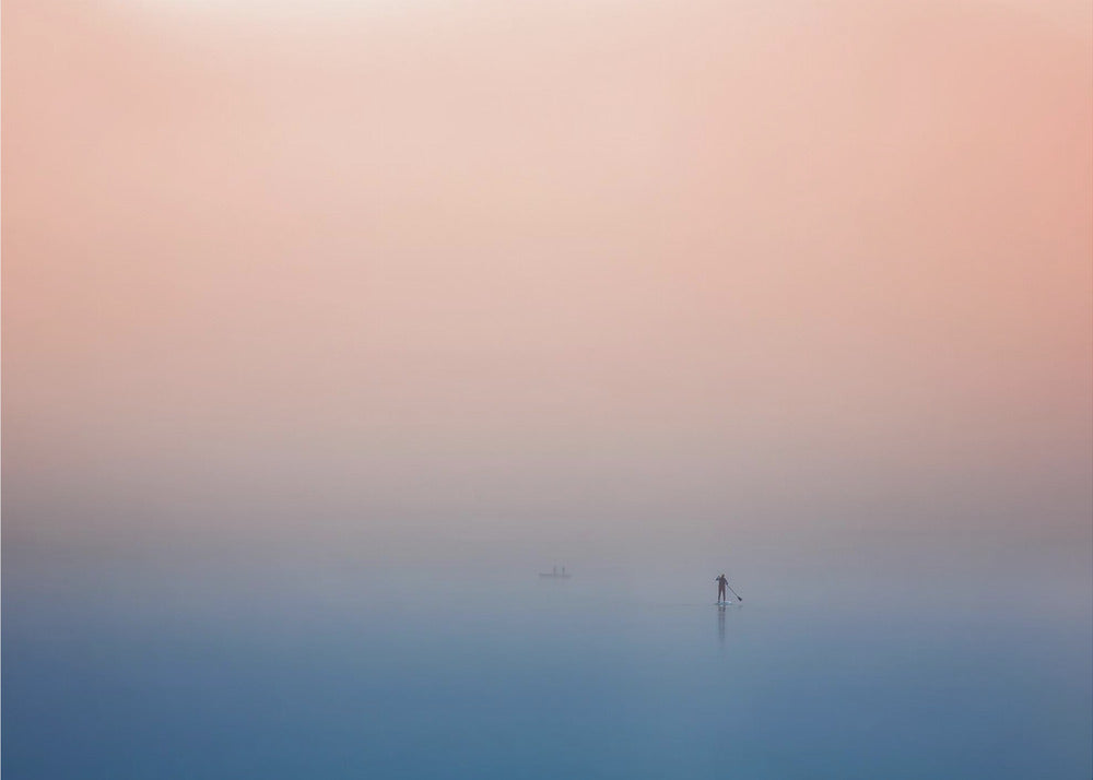 A minimalist framed photograph of a solitary person on a stand-up paddleboard, silhouetted against a vast, calm expanse of water under a hazy, gradient sky that shifts from soft pink to deep blue. Wall Art