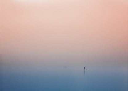 A minimalist framed photograph of a solitary person on a stand-up paddleboard, silhouetted against a vast, calm expanse of water under a hazy, gradient sky that shifts from soft pink to deep blue. Wall Art