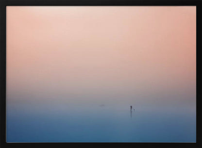 A minimalist framed photograph of a solitary person on a stand-up paddleboard, silhouetted against a vast, calm expanse of water under a hazy, gradient sky that shifts from soft pink to deep blue. Wall Art