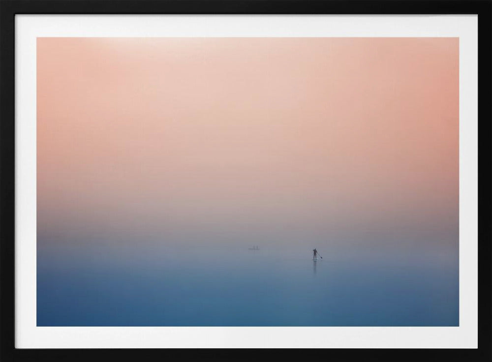 A minimalist framed photograph of a solitary person on a stand-up paddleboard, silhouetted against a vast, calm expanse of water under a hazy, gradient sky that shifts from soft pink to deep blue. Wall Art