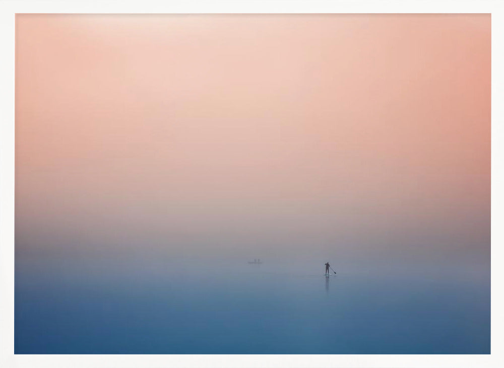 A minimalist framed photograph of a solitary person on a stand-up paddleboard, silhouetted against a vast, calm expanse of water under a hazy, gradient sky that shifts from soft pink to deep blue. Wall Art