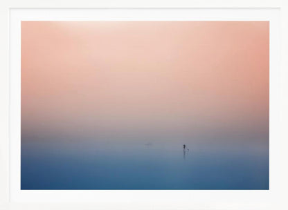 A minimalist framed photograph of a solitary person on a stand-up paddleboard, silhouetted against a vast, calm expanse of water under a hazy, gradient sky that shifts from soft pink to deep blue. Wall Art