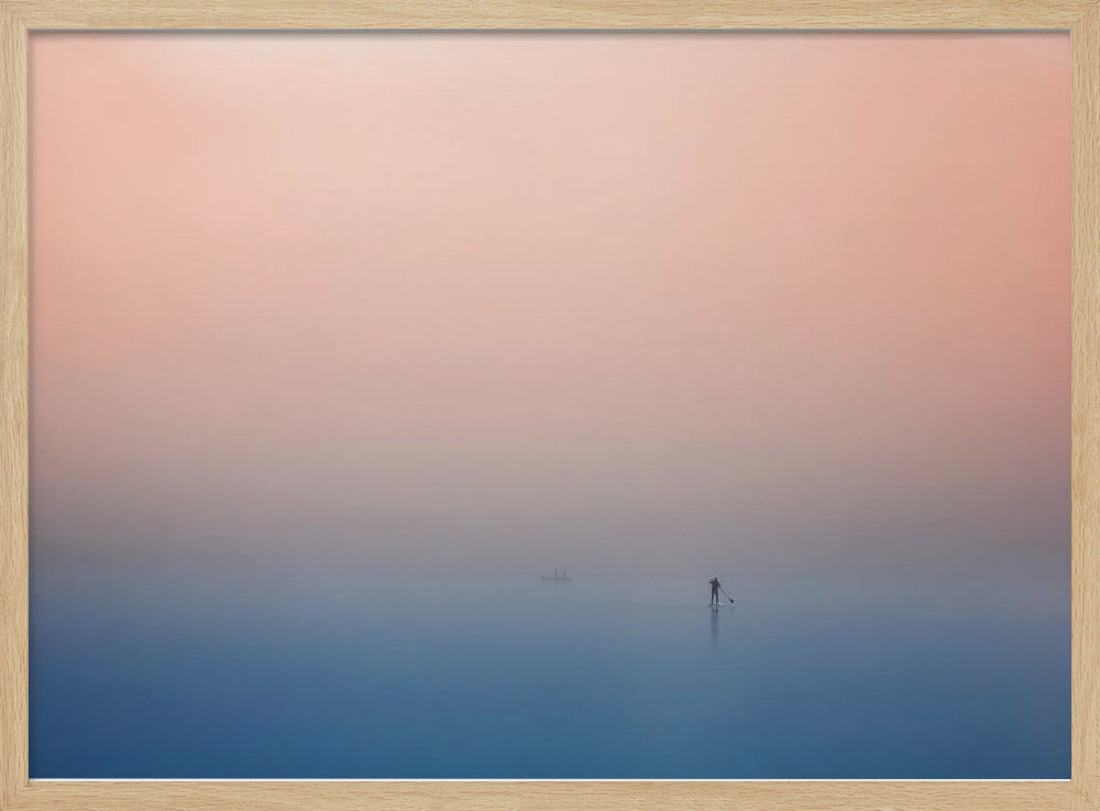 A minimalist framed photograph of a solitary person on a stand-up paddleboard, silhouetted against a vast, calm expanse of water under a hazy, gradient sky that shifts from soft pink to deep blue. Wall Art