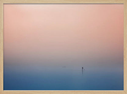 A minimalist framed photograph of a solitary person on a stand-up paddleboard, silhouetted against a vast, calm expanse of water under a hazy, gradient sky that shifts from soft pink to deep blue. Wall Art