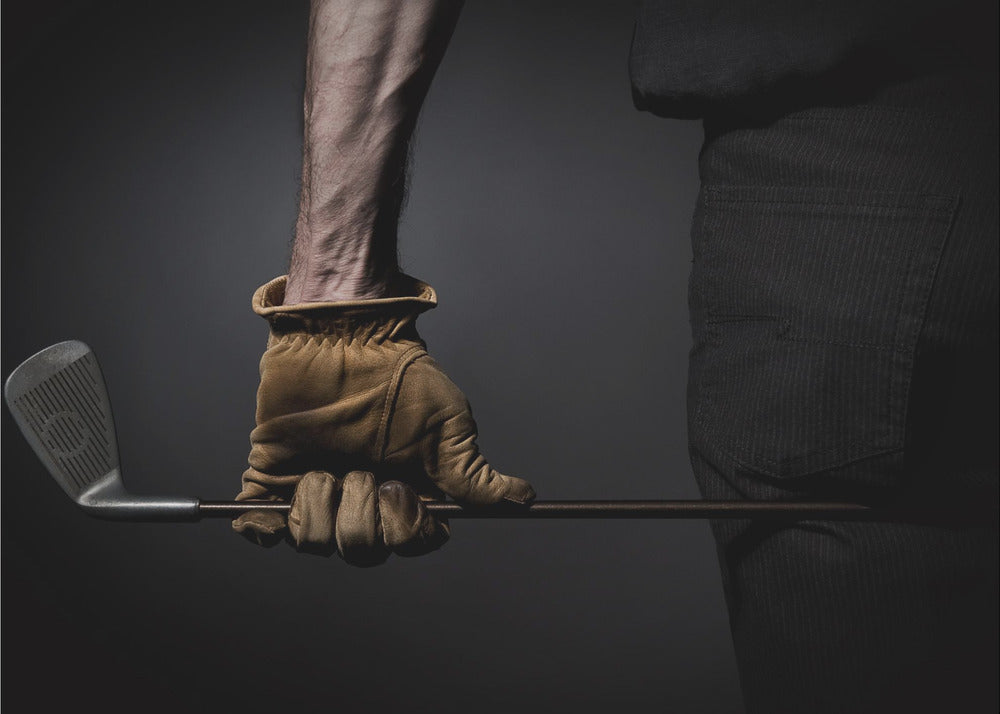 A dramatic close-up of a person's muscular forearm and hand wearing a tan leather glove, holding a golf iron horizontally against a dark background, framed in silver. Wall Art