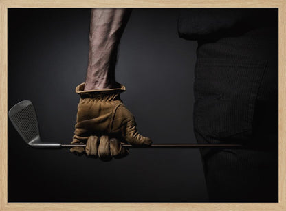 A dramatic close-up of a person's muscular forearm and hand wearing a tan leather glove, holding a golf iron horizontally against a dark background, framed in silver. Wall Art