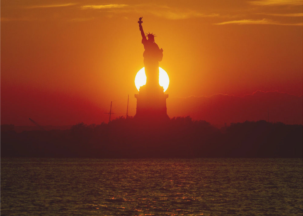 A powerful silhouette of the Statue of Liberty against a fiery orange and red sunset. The sun is perfectly positioned behind the statue's base, creating a brilliant, glowing orb of light. The dark waters in the foreground reflect the vibrant colors of the sky. The photograph is presented within a silver-colored frame. Artwork