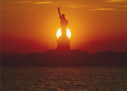 A powerful silhouette of the Statue of Liberty against a fiery orange and red sunset. The sun is perfectly positioned behind the statue's base, creating a brilliant, glowing orb of light. The dark waters in the foreground reflect the vibrant colors of the sky. The photograph is presented within a silver-colored frame. Artwork