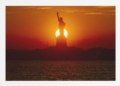A powerful silhouette of the Statue of Liberty against a fiery orange and red sunset. The sun is perfectly positioned behind the statue's base, creating a brilliant, glowing orb of light. The dark waters in the foreground reflect the vibrant colors of the sky. The photograph is presented within a silver-colored frame. Artwork