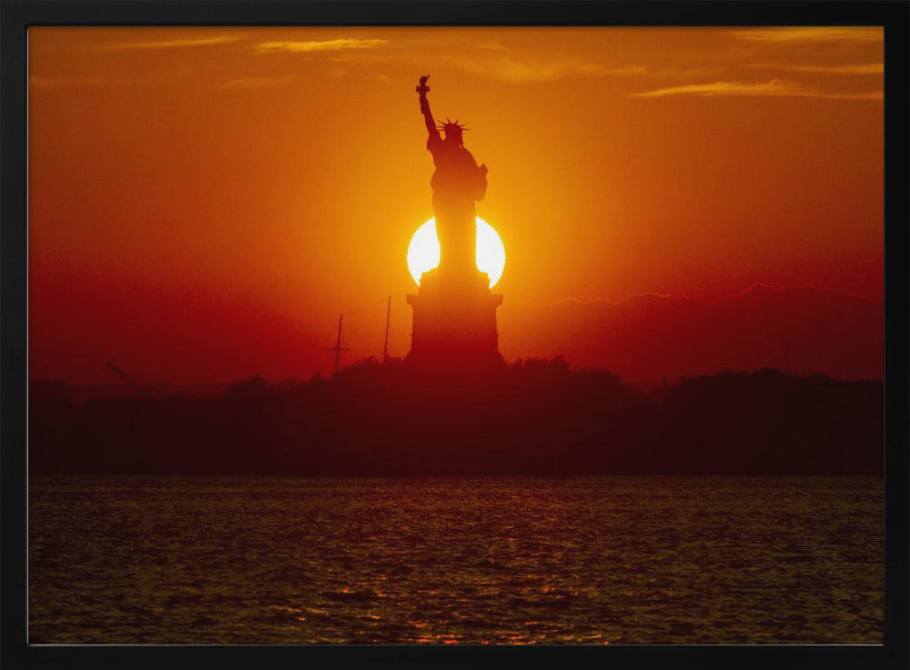 A powerful silhouette of the Statue of Liberty against a fiery orange and red sunset. The sun is perfectly positioned behind the statue's base, creating a brilliant, glowing orb of light. The dark waters in the foreground reflect the vibrant colors of the sky. The photograph is presented within a silver-colored frame. Artwork