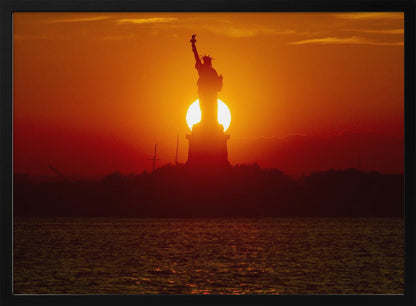 A powerful silhouette of the Statue of Liberty against a fiery orange and red sunset. The sun is perfectly positioned behind the statue's base, creating a brilliant, glowing orb of light. The dark waters in the foreground reflect the vibrant colors of the sky. The photograph is presented within a silver-colored frame. Artwork
