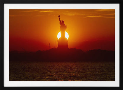 A powerful silhouette of the Statue of Liberty against a fiery orange and red sunset. The sun is perfectly positioned behind the statue's base, creating a brilliant, glowing orb of light. The dark waters in the foreground reflect the vibrant colors of the sky. The photograph is presented within a silver-colored frame. Artwork