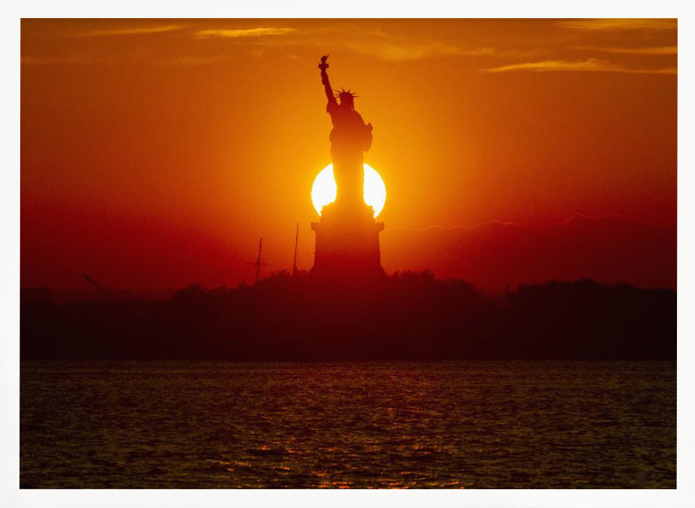 A powerful silhouette of the Statue of Liberty against a fiery orange and red sunset. The sun is perfectly positioned behind the statue's base, creating a brilliant, glowing orb of light. The dark waters in the foreground reflect the vibrant colors of the sky. The photograph is presented within a silver-colored frame. Artwork