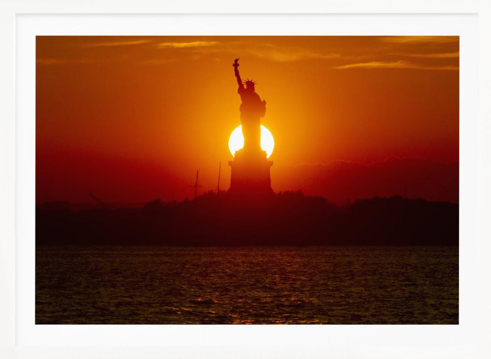 A powerful silhouette of the Statue of Liberty against a fiery orange and red sunset. The sun is perfectly positioned behind the statue's base, creating a brilliant, glowing orb of light. The dark waters in the foreground reflect the vibrant colors of the sky. The photograph is presented within a silver-colored frame. Artwork