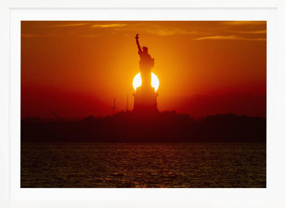 A powerful silhouette of the Statue of Liberty against a fiery orange and red sunset. The sun is perfectly positioned behind the statue's base, creating a brilliant, glowing orb of light. The dark waters in the foreground reflect the vibrant colors of the sky. The photograph is presented within a silver-colored frame. Artwork