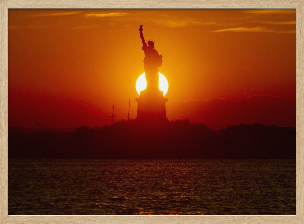 A powerful silhouette of the Statue of Liberty against a fiery orange and red sunset. The sun is perfectly positioned behind the statue's base, creating a brilliant, glowing orb of light. The dark waters in the foreground reflect the vibrant colors of the sky. The photograph is presented within a silver-colored frame. Artwork