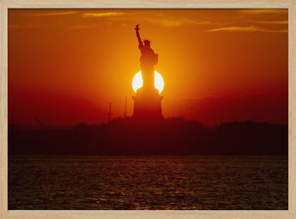 A powerful silhouette of the Statue of Liberty against a fiery orange and red sunset. The sun is perfectly positioned behind the statue's base, creating a brilliant, glowing orb of light. The dark waters in the foreground reflect the vibrant colors of the sky. The photograph is presented within a silver-colored frame. Artwork
