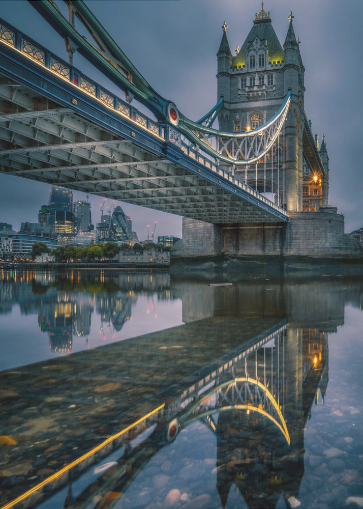 A dramatic, low-angle photograph of London's Tower Bridge at dusk. The iconic bridge is illuminated with warm yellow lights against a dark blue, cloudy sky. The calm River Thames in the foreground creates a stunning, clear reflection of the bridge and the distant city skyline. Print