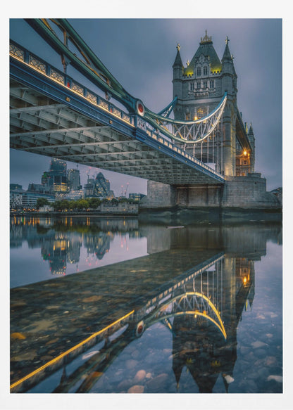 A dramatic, low-angle photograph of London's Tower Bridge at dusk. The iconic bridge is illuminated with warm yellow lights against a dark blue, cloudy sky. The calm River Thames in the foreground creates a stunning, clear reflection of the bridge and the distant city skyline. Print