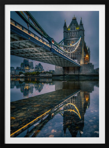 A dramatic, low-angle photograph of London's Tower Bridge at dusk. The iconic bridge is illuminated with warm yellow lights against a dark blue, cloudy sky. The calm River Thames in the foreground creates a stunning, clear reflection of the bridge and the distant city skyline. Print