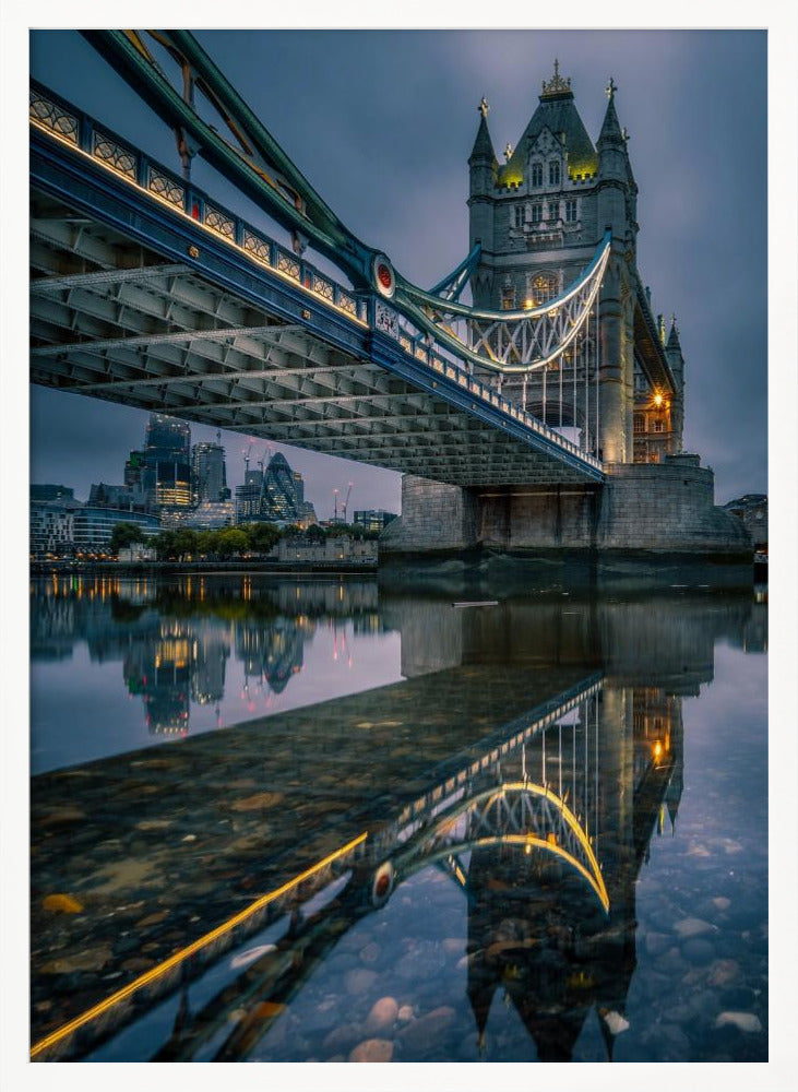 A dramatic, low-angle photograph of London's Tower Bridge at dusk. The iconic bridge is illuminated with warm yellow lights against a dark blue, cloudy sky. The calm River Thames in the foreground creates a stunning, clear reflection of the bridge and the distant city skyline. Print