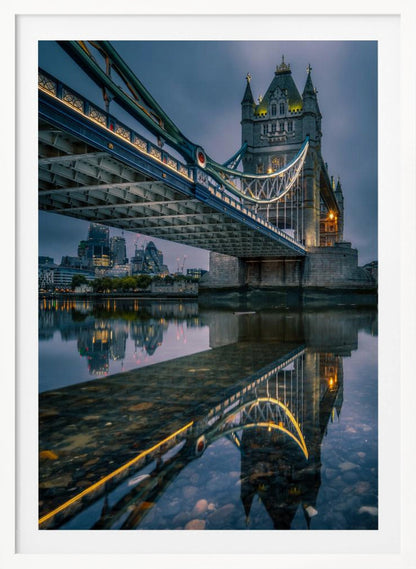 A dramatic, low-angle photograph of London's Tower Bridge at dusk. The iconic bridge is illuminated with warm yellow lights against a dark blue, cloudy sky. The calm River Thames in the foreground creates a stunning, clear reflection of the bridge and the distant city skyline. Print