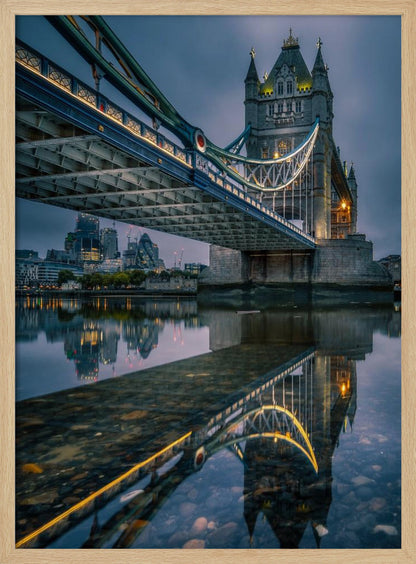 A dramatic, low-angle photograph of London's Tower Bridge at dusk. The iconic bridge is illuminated with warm yellow lights against a dark blue, cloudy sky. The calm River Thames in the foreground creates a stunning, clear reflection of the bridge and the distant city skyline. Print