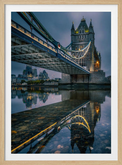 A dramatic, low-angle photograph of London's Tower Bridge at dusk. The iconic bridge is illuminated with warm yellow lights against a dark blue, cloudy sky. The calm River Thames in the foreground creates a stunning, clear reflection of the bridge and the distant city skyline. Print