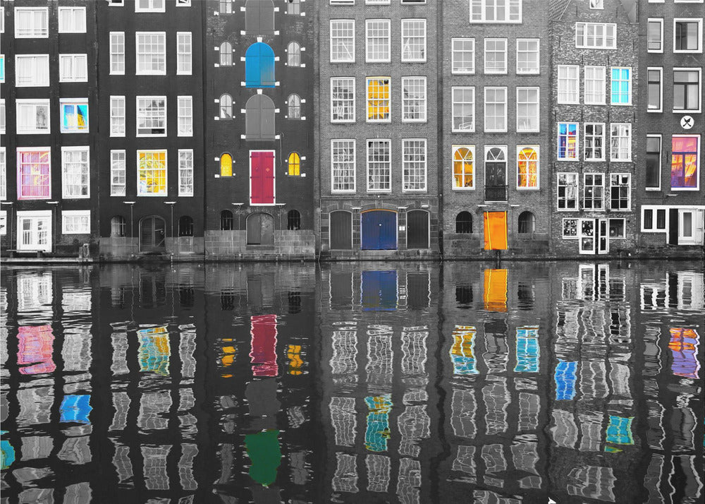 A framed black and white photograph of canal houses in Amsterdam, with selective coloring highlighting windows and doors in vibrant shades of red, blue, yellow, and pink. The colorful architectural details are mirrored in the rippling water of the canal below. Artwork