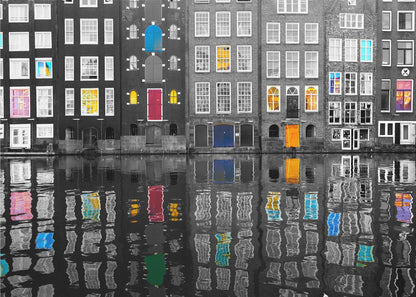 A framed black and white photograph of canal houses in Amsterdam, with selective coloring highlighting windows and doors in vibrant shades of red, blue, yellow, and pink. The colorful architectural details are mirrored in the rippling water of the canal below. Artwork