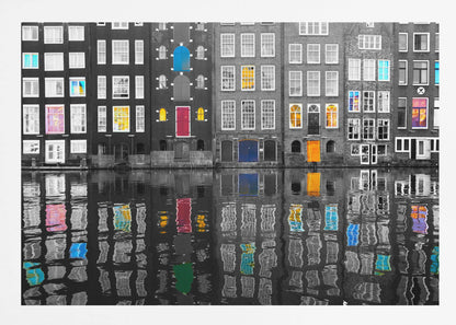 A framed black and white photograph of canal houses in Amsterdam, with selective coloring highlighting windows and doors in vibrant shades of red, blue, yellow, and pink. The colorful architectural details are mirrored in the rippling water of the canal below. Artwork