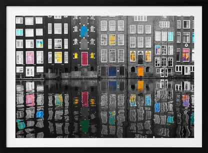 A framed black and white photograph of canal houses in Amsterdam, with selective coloring highlighting windows and doors in vibrant shades of red, blue, yellow, and pink. The colorful architectural details are mirrored in the rippling water of the canal below. Artwork