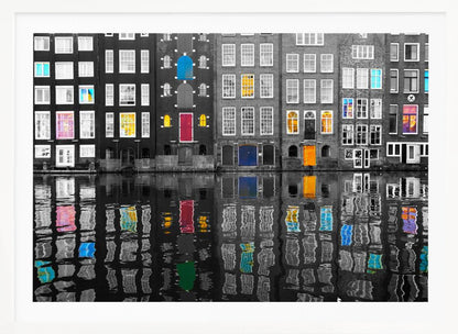 A framed black and white photograph of canal houses in Amsterdam, with selective coloring highlighting windows and doors in vibrant shades of red, blue, yellow, and pink. The colorful architectural details are mirrored in the rippling water of the canal below. Artwork
