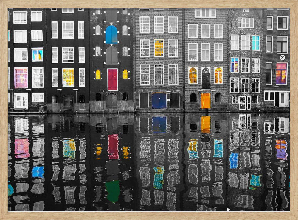 A framed black and white photograph of canal houses in Amsterdam, with selective coloring highlighting windows and doors in vibrant shades of red, blue, yellow, and pink. The colorful architectural details are mirrored in the rippling water of the canal below. Artwork