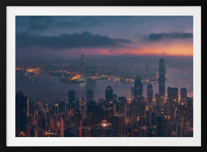 An aerial photograph of a sprawling city skyline at dusk, likely Hong Kong, with countless illuminated skyscrapers. The sky above transitions from a warm orange glow at the horizon to a deep purple, with dark clouds overhead. The city lights cast a golden reflection on the water of the harbor. The entire image is presented within a silver-colored frame. Poster