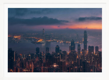 An aerial photograph of a sprawling city skyline at dusk, likely Hong Kong, with countless illuminated skyscrapers. The sky above transitions from a warm orange glow at the horizon to a deep purple, with dark clouds overhead. The city lights cast a golden reflection on the water of the harbor. The entire image is presented within a silver-colored frame. Poster