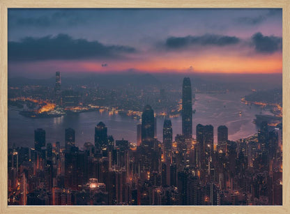 An aerial photograph of a sprawling city skyline at dusk, likely Hong Kong, with countless illuminated skyscrapers. The sky above transitions from a warm orange glow at the horizon to a deep purple, with dark clouds overhead. The city lights cast a golden reflection on the water of the harbor. The entire image is presented within a silver-colored frame. Poster